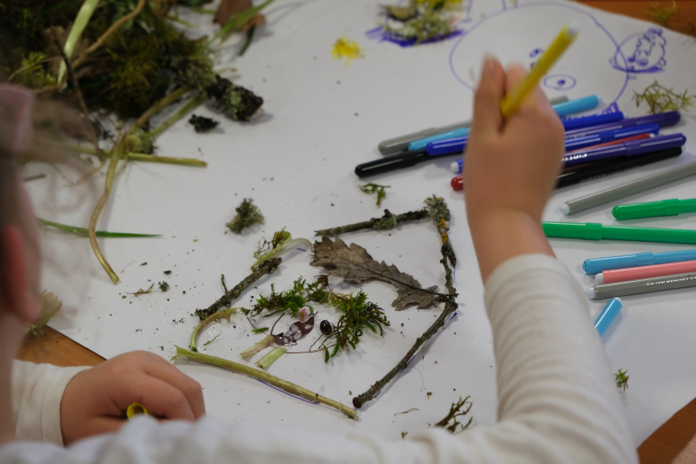 Un bras d'enfant dessine une maison avec des mousses et des branchages récupérés dans la nature